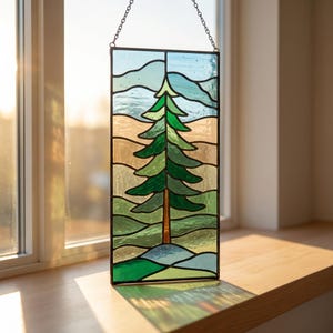 May include: A rectangular stained glass panel featuring a central evergreen tree with green foliage and a brown trunk. The background consists of horizontal wavy bands in shades of light blue, tan, and green. The panel is framed in dark metal.