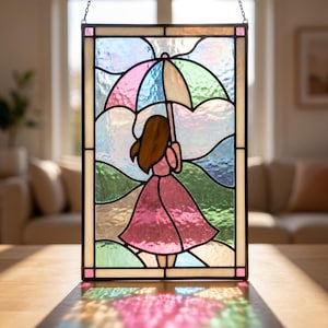 May include: A rectangular stained glass panel featuring a person with brown hair wearing a pink dress and holding a multi-colored umbrella. The glass uses shades of pink, blue, green, and white with a dark metal frame and a hanging chain.