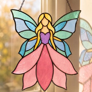 May include: A stained glass suncatcher shaped like a fairy with yellow hair, a purple bodice, and a pink petal-like skirt. The wings feature translucent panels of teal and light blue glass, all held together by dark metal solder lines and a chain.