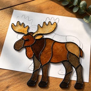 May include: A stained glass moose figure featuring various shades of brown and amber glass pieces joined by dark metal solder lines, placed over a white paper pattern template with numbered sections for assembly and construction.