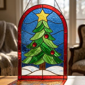May include: An arched stained glass panel featuring a green Christmas tree with red ornaments and a yellow star topper. The tree is set against a textured blue background with a white snowy base, all framed by a red border and suspended by a metal chain.