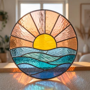 May include: A circular stained glass suncatcher featuring a bright yellow sun rising over stylized ocean waves. The sky is composed of textured peach and orange glass segments, while the water consists of layered blue and teal glass panels.