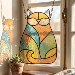 May include: A stained glass suncatcher shaped like a sitting cat, featuring segmented panels of iridescent orange, yellow, teal, and green glass held together by dark metal solder lines, suspended by a thin metal chain in front of a window.