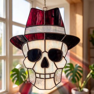 May include: A stained glass suncatcher shaped like a human skull wearing a fedora hat. The skull is constructed from off-white textured glass with black glass eye sockets and nose. The hat features deep red glass panels and a white glass band.