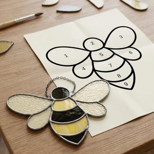 Puede incluir: Un atrapasoles de vidriera con forma de abeja, con paneles de vidrio texturizado amarillo y negro y una cadena de metal, junto a una plantilla de papel blanco con un dibujo lineal numerado de una abeja con secciones del uno al nueve.