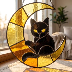 May include: A stained glass suncatcher featuring a black cat sitting on a yellow crescent moon. The cat has bright yellow eyes and is constructed from dark glass pieces. The moon is made of textured yellow glass segments held by metal cames.