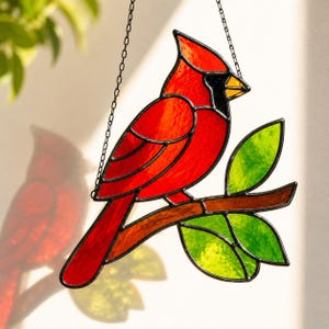 May include: A stained glass suncatcher featuring a bright red cardinal bird with black facial markings perched on a brown branch with vibrant green leaves. The piece is constructed with dark metal lead lines and hangs from a thin black metal chain.