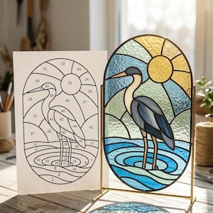 May include: An oval stained glass panel featuring a heron standing in water with a yellow sun in the background, supported by a gold metal stand. Beside it is a white paper template with numbered sections corresponding to the glass piece design.
