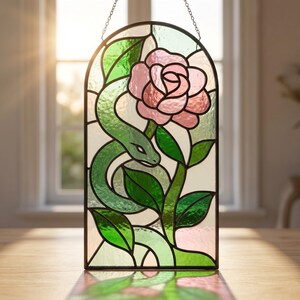 Può includere: Un pannello in vetro colorato ad arco raffigurante una rosa rosa centrale con foglie verdi e un serpente verde avvolto attorno al gambo. Il design è composto da vari segmenti di vetro tenuti insieme da profili metallici scuri.