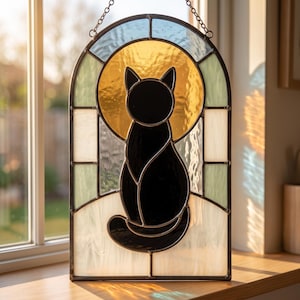 Könnte beinhalten: Ein bogenförmiger Buntglas-Sonnenfänger mit der Silhouette einer schwarzen Katze vor einem kreisförmigen, goldgelben Hintergrund. Die umliegenden Paneele bestehen aus strukturierten weißen und hellblauen Glasstücken, die durch dunkle Bleilinien verbunden sind.