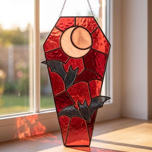 May include: A coffin-shaped stained glass suncatcher featuring a deep red background with a pale crescent moon at the top and two dark grey bats in flight. The piece is framed in black metal solder and hangs from a silver-toned metal chain.