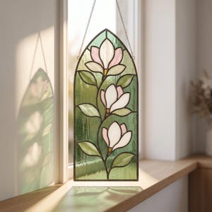 Puede incluir: Un panel de vidriera arqueado que muestra tres flores de magnolia blancas y rosa pálido con hojas verdes sobre un fondo verde texturizado. La pieza está enmarcada en metal oscuro y suspendida por una fina cadena plateada desde el marco superior.
