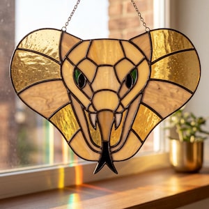 May include: A stained glass suncatcher shaped like a cobra head with a wide hood. The piece features cream and amber-coloured glass segments joined by dark metal solder lines, with two small green glass eyes and a black glass forked tongue.
