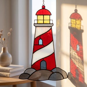 May include: A stained glass lighthouse suncatcher featuring a red and white spiral pattern, a yellow lantern room, and a gray rock base. The piece is constructed with dark metal solder lines and hangs from a small chain, casting a colorful light reflection.