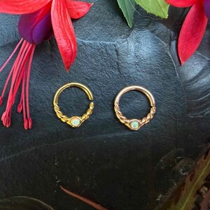 Pode incluir: Dois anéis circulares com uma textura de metal trançado e uma pequena pedra de opala iridescente engastada na base. Os anéis são exibidos sobre uma superfície escura e texturizada, emoldurados por pétalas de flores fúcsia rosa e violeta.