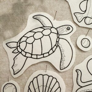 May include: A white fabric patch featuring a black embroidered outline of a sea turtle with a patterned shell. The patch is secured with a thin metal pin. Additional embroidered fabric shapes of a shell and bubbles are visible nearby.