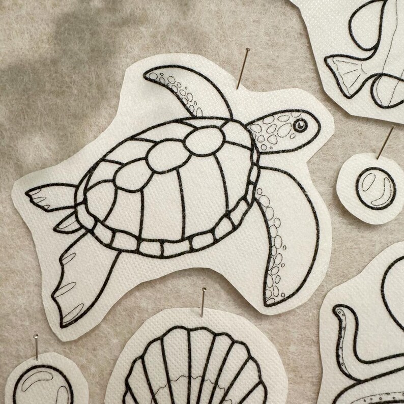 May include: A white fabric patch featuring a black embroidered outline of a sea turtle with a patterned shell. The patch is secured with a thin metal pin. Additional embroidered fabric shapes of a shell and bubbles are visible nearby.
