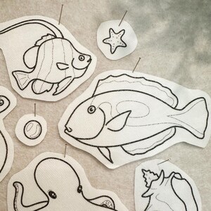 May include: A collection of white fabric patches featuring black line art illustrations of various sea creatures including two different fish, an octopus, a starfish, a seashell, and a bubble, each pinned with a small metal sewing pin.