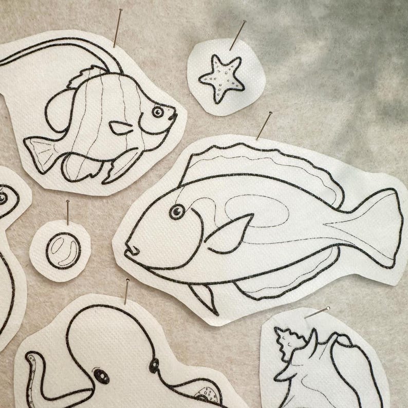 May include: A collection of white fabric patches featuring black line art illustrations of various sea creatures including two different fish, an octopus, a starfish, a seashell, and a bubble, each pinned with a small metal sewing pin.