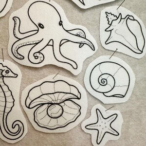May include: A collection of white fabric patches featuring black ink line art illustrations of marine life, including an octopus, a seahorse, a seashell, a spiral snail shell, a starfish, and an open clam shell containing a single pearl.