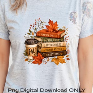 Op de afbeelding: Een lichtgrijs gemêleerd T-shirt met een afbeelding van een stapel boeken met de titels Pumpkin Spice, Crisp Air, Autumn Stories, Sweater Weather en Cozy Days, naast een koffiebeker met het opschrift Fall, omringd door oranje en rode herfstbladeren.