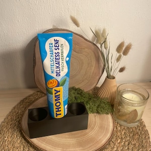 Könnte beinhalten: Eine blau-gelbe Tube Thomy Mittelscharfer Delikatess Senf mit einer Abbildung einer Senfblüte. Die Tube steht aufrecht in einem schwarzen, rechteckigen Halter mit zwei runden Aussparungen auf einer hölzernen Oberfläche.