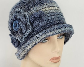 May include: A blue cloche hat with a floral accent. The hat is made of a chunky knit material in shades of blue and gray. The brim is slightly turned up, and a large flower is attached to the side.