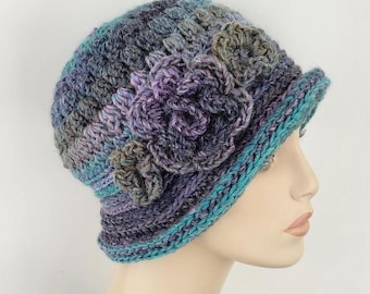 May include: A handmade crochet cloche hat in shades of blue, purple, and green. The hat features a decorative flower on the side and a turquoise brim. The hat is displayed on a mannequin head.