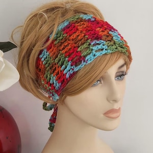 May include: A colorful, crocheted headband in shades of red, orange, green, and blue. The headband is displayed on a mannequin head with blonde hair. The headband is tied at the back.