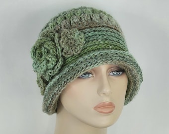 May include: A crochet cloche hat in variegated shades of sage, olive, and forest green. The hat features a textured stitch pattern and is adorned with two large, layered crochet flowers on the side, creating a dimensional and floral aesthetic.