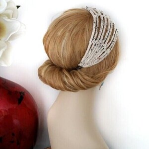 May include: A cream-colored, beaded headband with a unique, open design is worn in the hair. The hair is styled in a low bun. A red vase with white flowers is in the background.