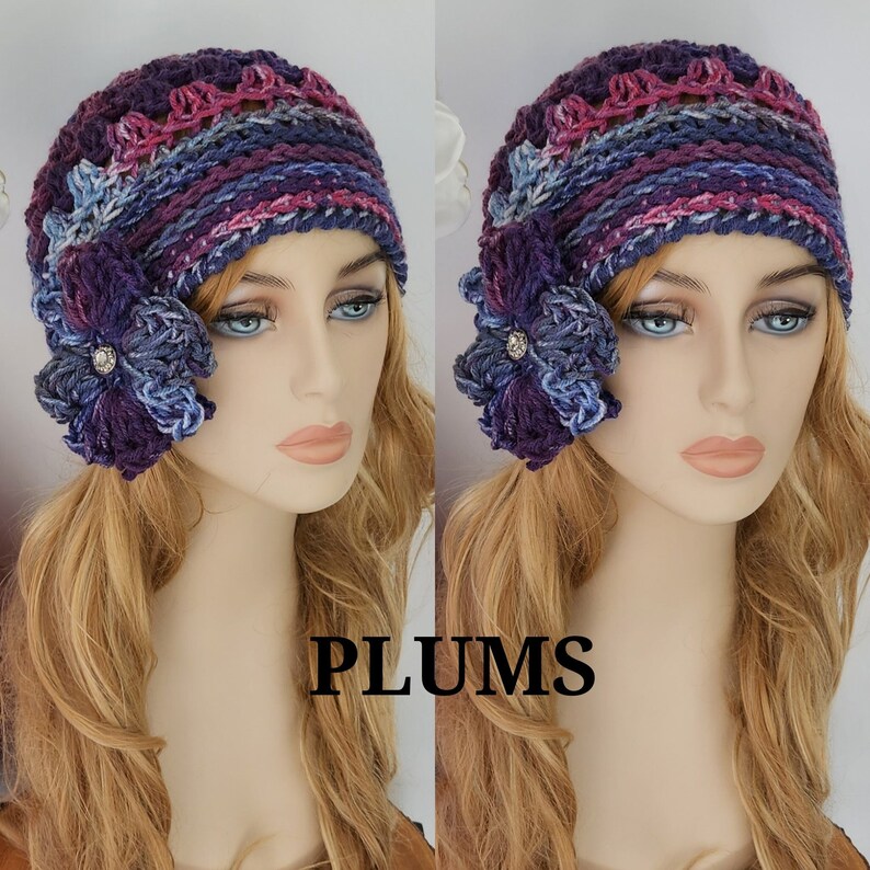 May include: A purple, blue, and pink crocheted hat with a matching flower embellishment. The hat is worn by a mannequin with long blonde hair. The word "PLUMS" is printed below the hat.