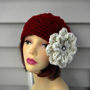 May include: A burgundy crochet beanie with a large, cream-colored flower accent. The flower is centered with a silver-colored button. The hat is displayed on a mannequin head.