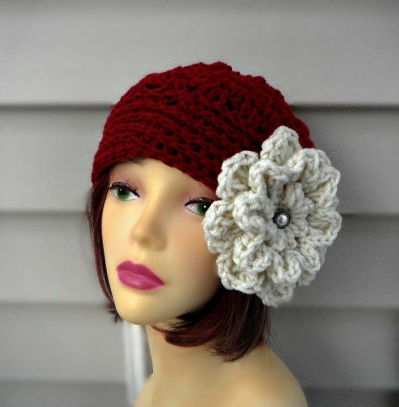 May include: A burgundy crochet beanie with a large, cream-colored flower accent. The flower is centered with a silver-colored button. The hat is displayed on a mannequin head.