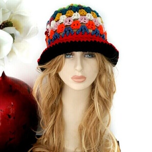 May include: A colorful crochet hat with a black brim. The hat features a patchwork design with various colors including red, orange, yellow, blue, and white. The hat is displayed on a mannequin head with long blonde hair.