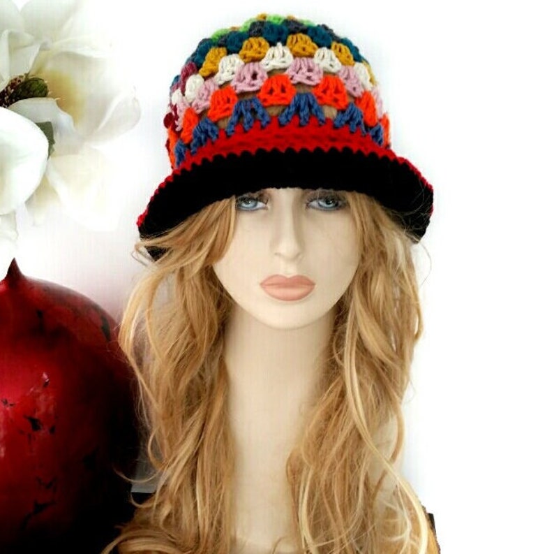 May include: A colorful crochet hat with a black brim. The hat features a patchwork design with various colors including red, orange, yellow, blue, and white. The hat is displayed on a mannequin head with long blonde hair.
