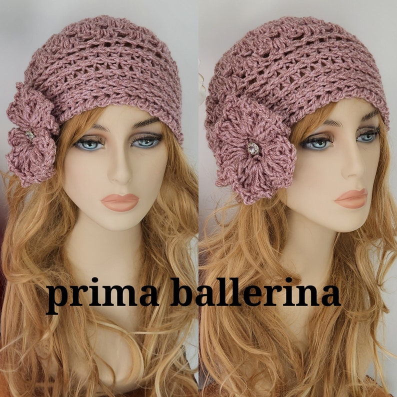 May include: A mauve crocheted beanie with a matching crocheted flower embellishment. The flower has a rhinestone in the center. The beanie is worn by a mannequin with long blonde hair. The text "prima ballerina" is printed below the mannequin.