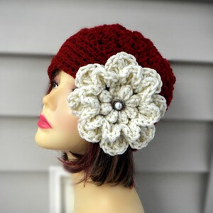 May include: A burgundy crochet hat with a large, cream-colored flower attached to the side. The flower has a pearl-like center. The hat is made of a textured yarn and is displayed on a mannequin head.