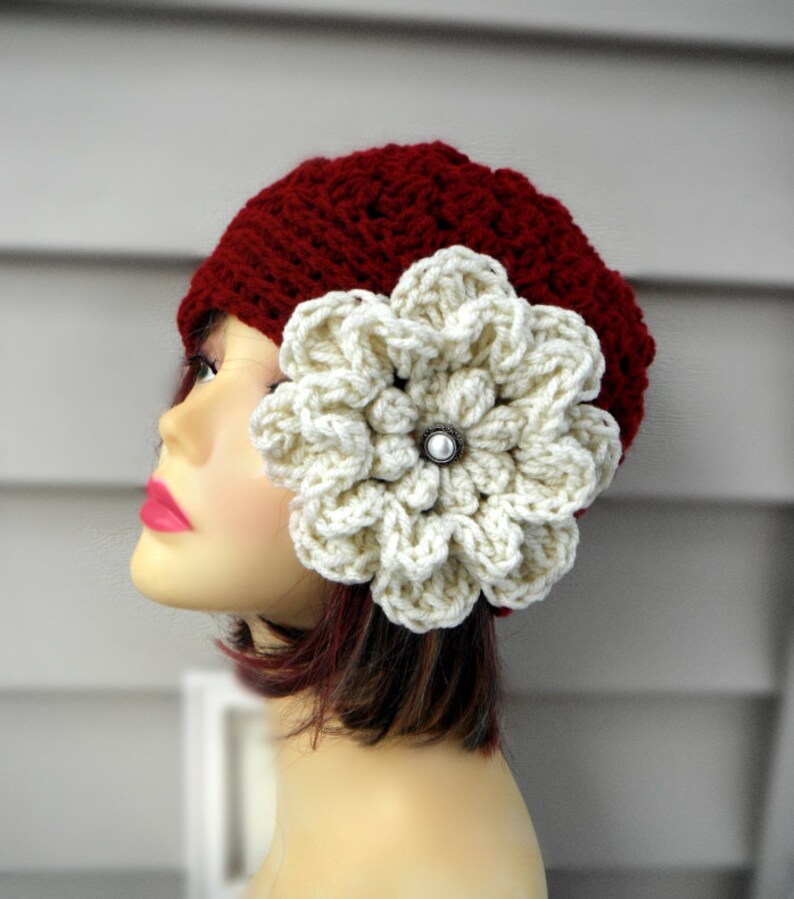 May include: A burgundy crochet hat with a large, cream-colored flower attached to the side. The flower has a pearl-like center. The hat is made of a textured yarn and is displayed on a mannequin head.