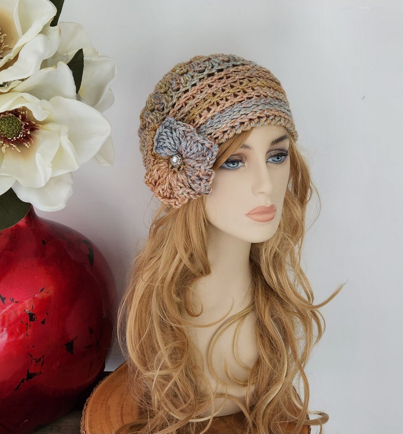 May include: A crocheted hat with a flower embellishment. The hat is made in a variety of shades of brown, pink, and blue. The flower is a light blue and pink color.