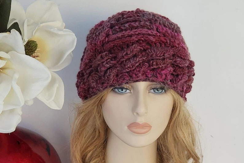 May include: A close-up of a knitted hat in shades of pink and burgundy. The hat has a textured, braided design and is worn by a mannequin with blonde hair.