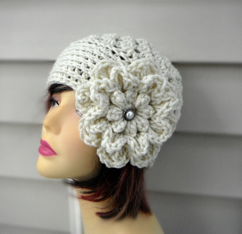 May include: A white crocheted beanie with a large white crocheted flower on the side. The flower has a pearl-like button in the center.
