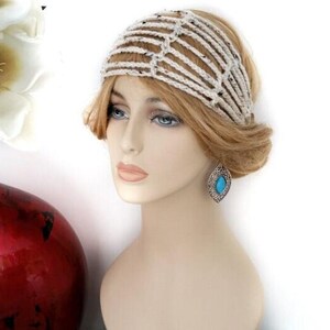 May include: A mannequin head displays a cream-colored crochet headband with black flecks. The headband is styled with a side bun and a turquoise teardrop earring. A red vase with white flowers is in the background.