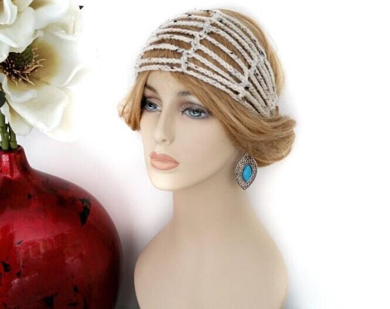 May include: A mannequin head displays a cream-colored crochet headband with black flecks. The headband is styled with a side bun and a turquoise teardrop earring. A red vase with white flowers is in the background.