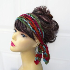 May include: A colorful crochet headband with a braided design. The headband features strands of red, green, blue, and orange yarn. The ends of the headband are attached to a silver hoop. The headband is worn on a mannequin head.