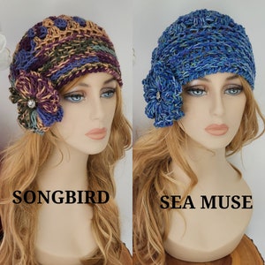 May include: Two crocheted hats with flower embellishments. The hat on the left is a multi-colored pattern with a purple flower. The hat on the right is blue with a blue flower. The hats are labeled "Songbird" and "Sea Muse".