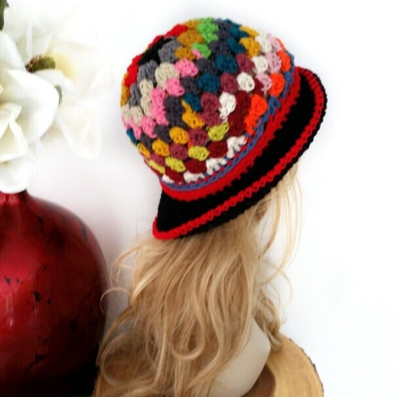 May include: A colorful crochet hat with a black brim and red and blue trim. The hat features a patchwork design with various colors, including pink, yellow, green, and blue. The hat is shown on a mannequin head with blonde hair.