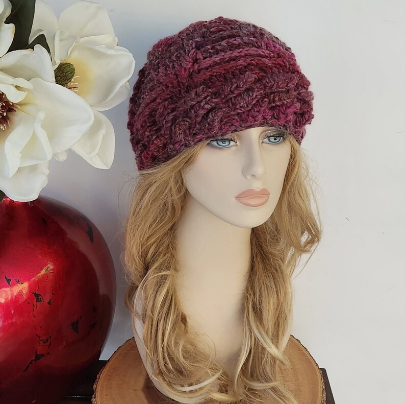 May include: A pink and purple knitted headband with a braided design. The headband is worn by a mannequin with long blonde hair.