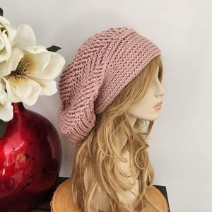 May include: A dusty rose slouchy beanie hat featuring a prominent herringbone knit pattern. The hat is crafted from a thick yarn and covers the top of the head with a relaxed fit, showcasing a textured stitch design throughout the entire surface.