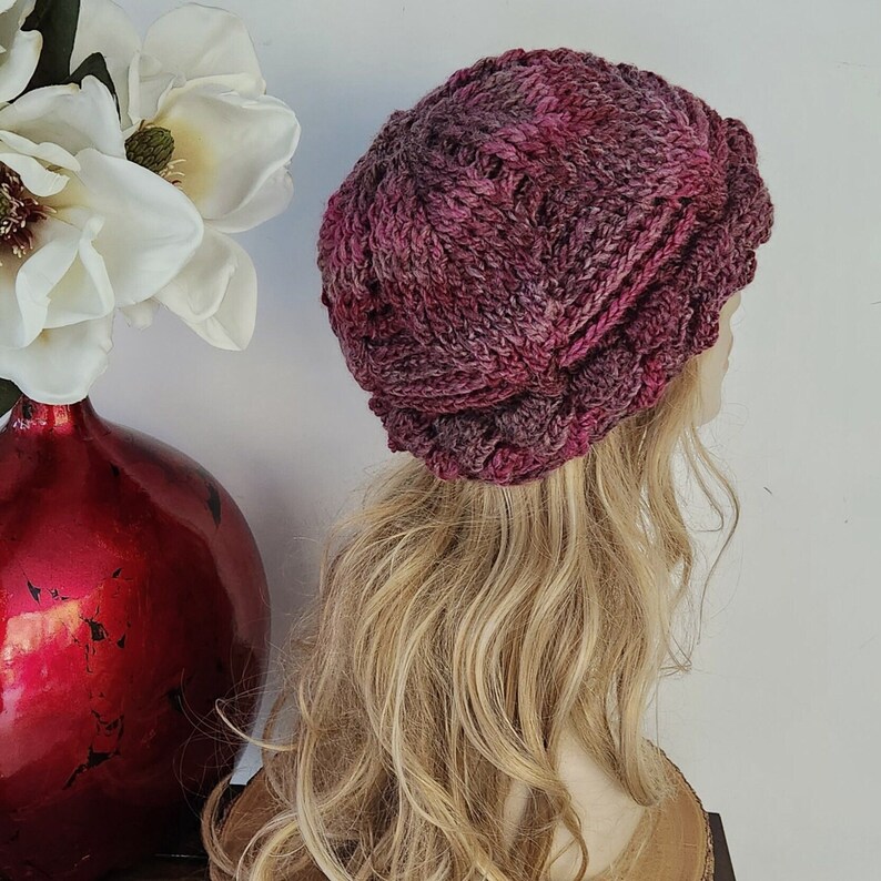 May include: A close-up of a knitted hat in shades of pink and purple. The hat has a textured, cable knit design and is shown on a mannequin with long blonde hair.